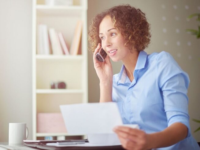 woman in blue shirt reading document while on a cordless landline phone